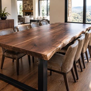 May include: A large rectangular dining table featuring a dark brown natural live edge wood slab top supported by two thick black metal U-shaped legs, accompanied by eight upholstered chairs with light brown seats and dark wooden frames.
