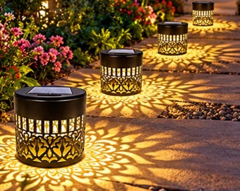 Op de afbeelding: Een reeks cilindervormige zwarte solartuinlampen met ingewikkelde uitgesneden patronen. Elke lamp heeft een zonnepaneel aan de bovenkant en straalt een warm licht uit dat gedetailleerde bloemvormige schaduwen op het stenen pad werpt.