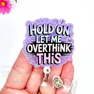 May include: A purple and white badge with the text "HOLD ON LET ME OVERTHINK THIS". The badge has a glittery border and a retractable reel clip. The badge is held in a hand.