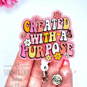 May include: A pink badge reel with the words "CREATED WITH A PURPOSE" in a retro font, decorated with flowers and a butterfly. The badge reel has a clear plastic backing and a metal clip.
