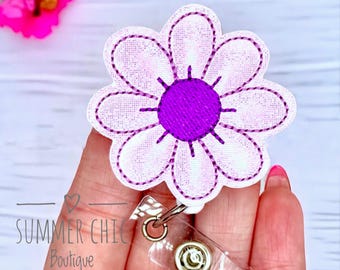 May include: A white and purple flower-shaped badge reel with a stitched design. The centre of the flower is a solid purple, and the petals are white with purple stitching. The badge reel has a clear plastic strap and a metal clip. The text "SUMMER CHIC Boutique" is visible.