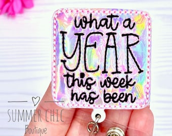 May include: A square badge reel with the text "what a YEAR this week has been" in black. The badge has a holographic background with a pink stitched border. The reel has a clear plastic strap and a metal clip.