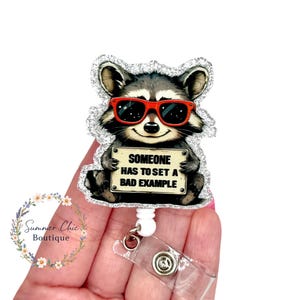 May include: A badge reel featuring a raccoon wearing red sunglasses, holding a sign that reads "SOMEONE HAS TO SET A BAD EXAMPLE." The raccoon is surrounded by a silver glitter border. The reel has a clear plastic clip.