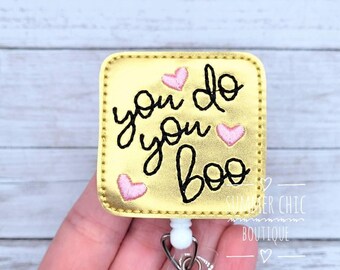 May include: A gold badge reel with the phrase "you do you boo" in black script, accented with pink heart shapes. The badge has a white retractable cord and a metal clip.