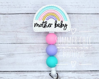 May include: A badge reel with a rainbow design and the words "mother baby" in black script. The rainbow features pastel colors. Below the rainbow are three colorful beads: pink, purple, and teal. The reel has a white cord and a silver clip.