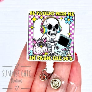May include: A white badge reel with a cartoon skeleton wearing headphones and holding a handheld game console. The badge has the text "BE PATIENT WITH ME I'M FROM THE 90'S". A pink Furby is also featured.