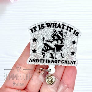 May include: A badge reel with a glittery design and the text "IT IS WHAT IT IS AND IT IS NOT GREAT". The image shows a raccoon in a chair. The reel has a clear plastic clip and a white retractable reel.