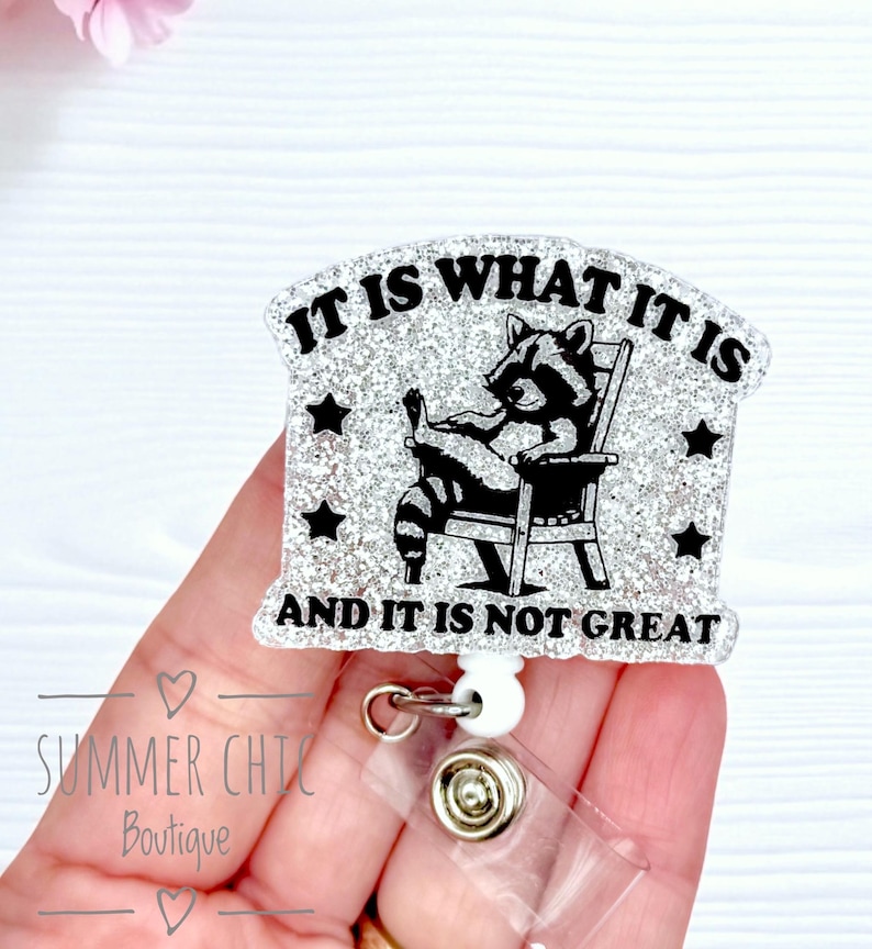 May include: A badge reel with a glittery design and the text "IT IS WHAT IT IS AND IT IS NOT GREAT". The image shows a raccoon in a chair. The reel has a clear plastic clip and a white retractable reel.