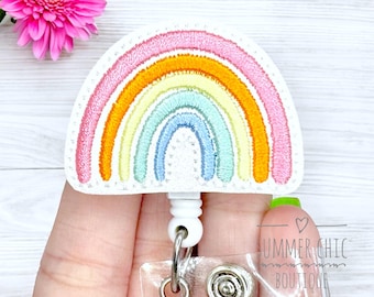 May include: A badge reel featuring a stitched rainbow design with a white border. The rainbow has pink, orange, yellow, green, and blue arcs. The reel has a white retractable cord and a clear plastic badge holder. A pink flower is in the background.