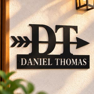 May include: A black metal wall sign featuring the large capital letters DT intersected by a horizontal arrow. Below the arrow, a rectangular plaque displays the name DANIEL THOMAS in bold, white, capitalized lettering.