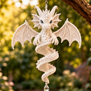 Peut inclure: Un ornement de dragon crocheté de couleur crème avec des motifs de points complexes, de grandes ailes, des cornes et une queue en spirale. La créature a des yeux bleu clair et est suspendue à un fil avec un cristal transparent en forme de goutte.