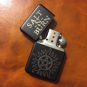 May include: Black lighter with a silver top and a black and white engraved symbol of a pentagram with flames around it. The top of the lighter has the words "Salt -n- Burn" engraved in gold.
