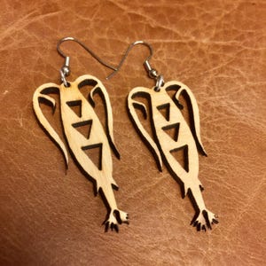 Puede incluir: Un par de pendientes de madera con un diseño único que presenta una figura estilizada con patrones triangulares. Los pendientes están hechos de madera marrón claro y tienen ganchos plateados.
