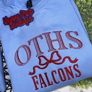 May include: A light blue sweatshirt with red embroidered text reading "OTHS Falcons." A red and blue glittery patch that says "Happy Crab Boutique" is attached to the top. The sweatshirt has a crew neck.