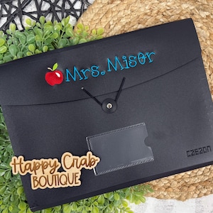 May include: Black document organizer with the name "Mrs. Miser" embroidered in blue, and a red apple graphic. The organizer has a clear pocket and a button closure. The brand name "caszon" is printed on the lower right.