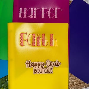 May include: A stack of colorful binders. The top binder is magenta with the name "Harper" embroidered in pink. Below is a yellow binder with the name "Faith" embroidered in orange and the text "Happy Crab Boutique". Other binders are green and blue.