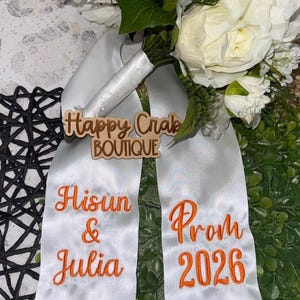 May include: A white floral bouquet with a satin ribbon. The ribbon has the text "Hisun & Julia" and "Prom 2026" embroidered in orange. A wooden sign with the text "Happy Crab Boutique" is attached.