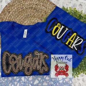 May include: A royal blue t-shirt with a black and gold glitter "Congrats" design and a blue, white, and yellow glitter "Cougars" design. The shirt is from Happy Crab Boutique.