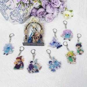 May include: A collection of acrylic keychains featuring various illustrated characters in wizard-like attire, including one larger standing acrylic display piece with an arched frame, all arranged on a white textured surface.