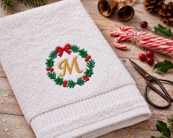 May include: A white terry cloth hand towel featuring an embroidered green holly wreath with red berries and a red bow, centered around a gold-stitched letter M. The towel has a textured border and a soft, absorbent fabric construction.