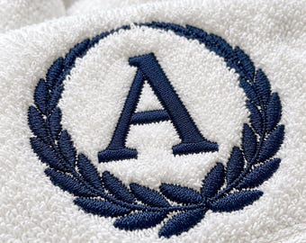 May include: A white terry cloth towel featuring a dark blue embroidered design. The embroidery consists of a capital letter A centered within a circular laurel wreath motif, showcasing detailed stitching on the textured fabric surface.