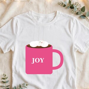 May include: A white short-sleeved t-shirt featuring a graphic of a bright pink mug filled with a dark brown beverage topped with white marshmallows. The word JOY is printed in white capital letters on the front of the pink mug.
