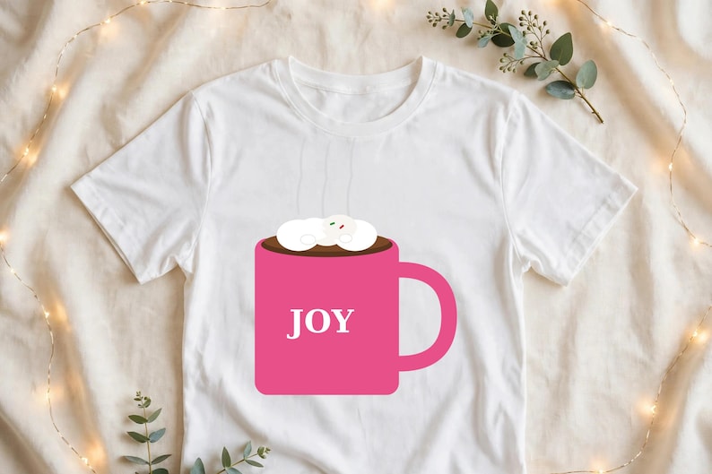 May include: A white short-sleeved t-shirt featuring a graphic of a bright pink mug filled with a dark brown beverage topped with white marshmallows. The word JOY is printed in white capital letters on the front of the pink mug.
