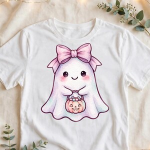May include: A white short-sleeved t-shirt featuring a central graphic of a small, smiling ghost with rosy cheeks. The ghost wears a large light pink bow on its head and holds a small orange jack-o'-lantern bucket filled with candy.