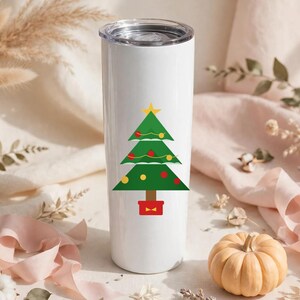 May include: A white cylindrical stainless steel tumbler featuring a graphic of a green Christmas tree decorated with red and yellow ornaments, a yellow star topper, and a brown trunk set inside a red square base with a clear plastic lid.
