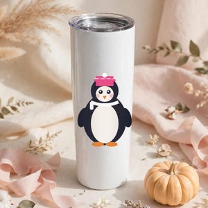 May include: A white cylindrical stainless steel tumbler featuring a graphic of a penguin wearing a pink hat and a white scarf. The penguin has a black body, a white belly, orange feet, and a small orange beak. The tumbler has a clear lid.