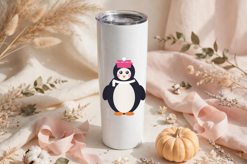 May include: A white cylindrical stainless steel tumbler featuring a graphic of a penguin wearing a pink hat and a white scarf. The penguin has a black body, a white belly, orange feet, and a small orange beak. The tumbler has a clear lid.
