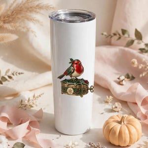 May include: A white cylindrical tumbler with a clear lid featuring a graphic of a red-breasted robin perched on a decorative, ornate, dark green and gold trinket box adorned with holly leaves and a small snowflake pattern.