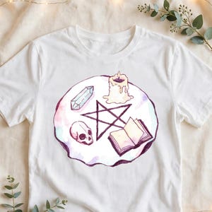 May include: A white short-sleeve t-shirt featuring a circular watercolor illustration. The design includes a dark purple pentagram, a human skull, a clear crystal, a lit candle, and an open book, all arranged on a soft pink and purple background.
