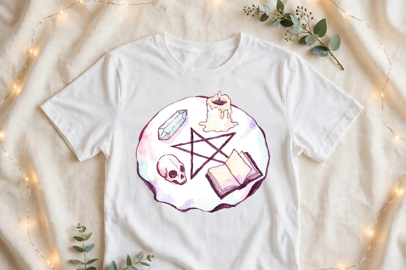 May include: A white short-sleeve t-shirt featuring a circular watercolor illustration. The design includes a dark purple pentagram, a human skull, a clear crystal, a lit candle, and an open book, all arranged on a soft pink and purple background.