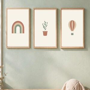 May include: Three framed wall art prints with light wood frames. The left print features a rainbow in muted red and green. The center print shows a green potted plant. The right print displays a hot air balloon in red and beige tones.