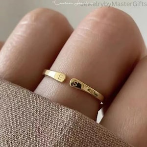 Puede incluir: Un anillo abierto de tono dorado con dos extremos planos y redondeados. Cada extremo está grabado con una letra mayúscula, la letra E en un lado y la letra S en el otro. La banda tiene un acabado liso y pulido en toda su superficie.