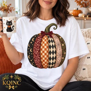 Peut inclure: Un t-shirt blanc orné d'un grand motif de citrouille aux motifs variés, surmonté d'un nœud rouge foncé. Les segments de la citrouille présentent des motifs célestes, floraux, à carreaux, en losanges et en toile d'araignée. Un mug assorti montre un chat noir.