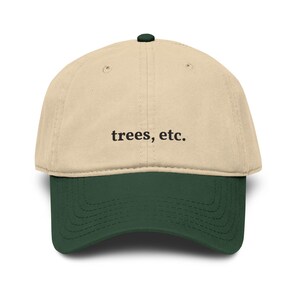 May include: A two-tone baseball cap featuring a beige crown and a dark green brim. The front of the cap displays the text "trees, etc." embroidered in black lettering. The design includes a small dark green button at the top center.