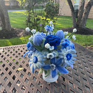 May include: A bouquet of handcrafted blue and white flowers made from pipe cleaners, featuring various shades of blue, including navy and light blue, arranged in a small white vase. The floral arrangement includes daisies, lilies, and small buds.