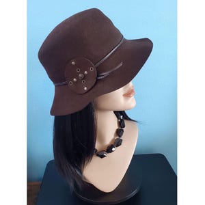 May include: A brown cloche hat with a decorative leather band and embellishment. The hat features a wide brim and is displayed on a mannequin head. The hat is a vintage style.