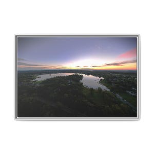 May include: A framed landscape wall art print featuring an aerial view of a lake surrounded by dense green trees at sunset. The sky transitions from deep purple to orange and pink hues above the horizon, reflecting light onto the water surface.