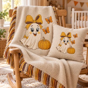 May include: A beige knit throw blanket and a matching square throw pillow, both featuring an embroidered design of a smiling ghost with a large yellow bow, a pumpkin, and orange butterflies. The blanket has a multi-coloured fringe edge.