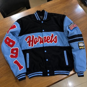 Könnte beinhalten: Eine schwarz-hellblaue Collegejacke mit dem Wort Hornets in roter Schreibschrift auf der Brust. Der rechte Ärmel zeigt die roten Zahlen 1981, während der linke Ärmel einen Delaware Hornets Aufnäher und einen kleineren HBUs Do More Aufnäher trägt.