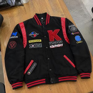 Puede incluir: Una chaqueta universitaria negra con mangas de efecto piel roja y puños y cuello de canalé a rayas. El frente presenta una gran letra K roja, el texto Kavinsky, Plasma, Renegade, Zombie y Outsider, junto con varios parches circulares.
