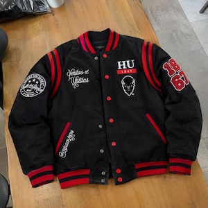 Könnte beinhalten: Eine schwarze Collegejacke mit rot gestreiften Rippbündchen an Ärmeln, Kragen und Saum. Die Vorderseite zeigt den Text HU 1867, eine Bison-Grafik und den Schriftzug Veritas et Utilitas. Der rechte Ärmel hat einen runden Howard University Aufnäher, der linke Ärmel zeigt 1867 in Rot.