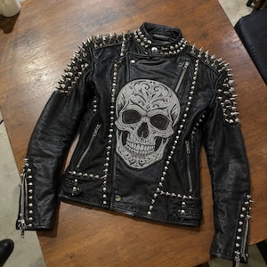 Könnte beinhalten: Eine schwarze Lederjacke mit silberfarbenen Metallnieten an den Schultern, am Kragen und an den Ärmeln. Auf der Vorderseite befindet sich ein großes, aufwendig gesticktes Totenkopfmotiv mit verzierten Mustern sowie silberne Reißverschlüsse.