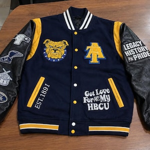 Puede incluir: Una chaqueta universitaria azul marino con detalles en amarillo y blanco, mangas de piel sintética negra, un parche de bulldog, un logotipo A&T y texto bordado que incluye EST. 1891, Got Love For My HBCU y Legacy History Pride con varios parches.