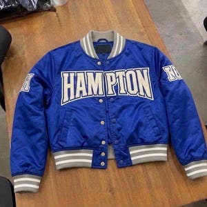Pode incluir: Uma jaqueta varsity azul royal com a palavra HAMPTON em letras grandes e brancas no peito. A jaqueta possui gola, punhos e bainha canelados com listras cinzas e brancas, além de botões de pressão prateados na parte frontal central.