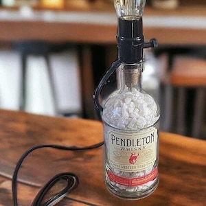 May include: A repurposed whiskey bottle lamp with a clear glass bulb and a black cord. The bottle is filled with white rocks and has a label that reads "Pendleton Whiskey True Western Tradition".