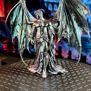 May include: A metallic, iridescent dark-toned figurine of a winged humanoid figure with horns, standing with large, expansive, textured wings spread wide. The figure wears a draped, flowing garment around the waist and lower body.